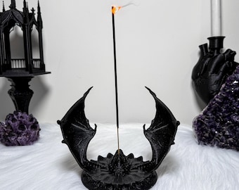 May include: Black dragon wing incense holder with a burning incense stick. The holder is on a white surface, with a black gothic structure and a purple crystal in the background. The incense stick emits a thin trail of smoke.