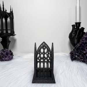 Puede incluir: Una pieza decorativa negra de estilo gótico, que se asemeja a una ventana de catedral, se encuentra sobre una superficie blanca. El intrincado diseño presenta ventanas arqueadas y soportes verticales. También se ven otras decoraciones de temática gótica.