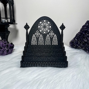 Pode incluir: Uma peça decorativa preta em estilo gótico que se assemelha a uma janela de catedral e uma escada. O design intrincado apresenta uma janela arqueada detalhada com um padrão circular e duas agulhas decorativas. A escada tem vários degraus e uma base decorativa.