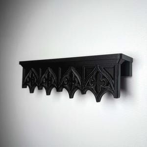 May include: A black, gothic-style shelf with intricate, repeating arched patterns. The shelf is mounted on a white wall, showcasing its detailed design and dark color. The shelf is approximately 12 inches long.