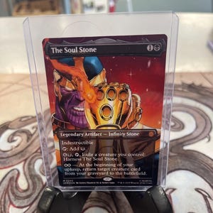 May include: A collectible card titled "The Soul Stone" featuring Thanos with the Infinity Gauntlet. The card is in a protective sleeve and held by a black stand. The card has a red and orange background.