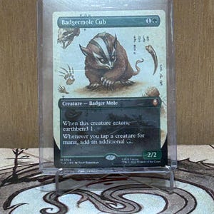 May include: A Magic: The Gathering card featuring a Badger Mole creature. The card is in a protective sleeve and displays the name "Badgermole Cub" at the top. The artwork depicts a brown and white badger-like creature. The card's text describes its abilities.