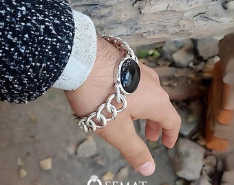 Puede incluir: Un brazalete de cadena de plata con una piedra ovalada negra. El brazalete está en la muñeca de una persona.