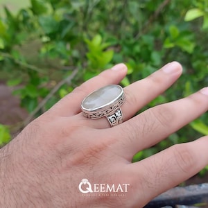 Pode incluir: Um anel de prata com uma gema branca ovalada engastada em um design floral detalhado.