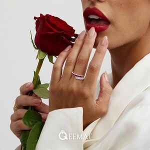 Könnte beinhalten: Nahaufnahme einer Hand mit einem Weißgoldring mit roten Edelsteinen und weißen Diamanten. Die Person hält eine rote Rose. Das Bild ist vor einem weißen Hintergrund aufgenommen.