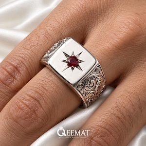 May include: A silver signet ring featuring a square face with a central red gemstone set within an engraved starburst pattern. The sides of the ring are decorated with intricate scrollwork engravings. The text QEEMAT is visible at the bottom.