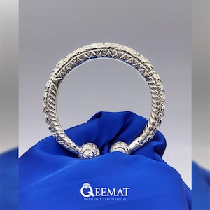 Puede incluir: Un brazalete de plata con diseños intrincados y una superficie texturizada. El brazalete está sobre un fondo azul.