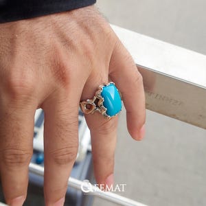Puede incluir: Un anillo de plata con una gran piedra turquesa rectangular. El anillo tiene un diseño calado y ornamentado en los laterales. La piedra turquesa es de un azul brillante. Es una joya.
