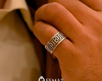 May include: A silver ring with a black Greek key design. The ring is worn on a person's finger.