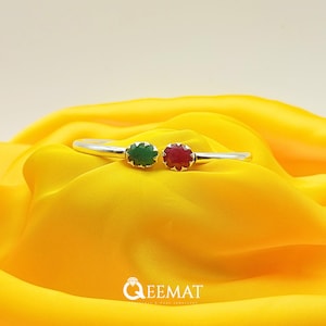 Può includere: Un braccialetto d'argento con un accento di pietra preziosa verde e rossa. Il braccialetto è su uno sfondo di tessuto giallo.
