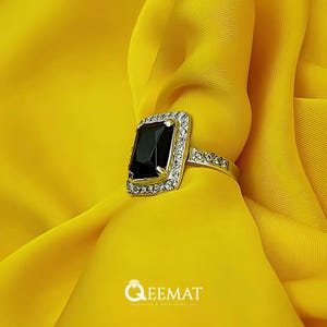 Könnte beinhalten: Ein goldfarbener Ring mit einem großen, rechteckigen schwarzen Edelstein. Der Edelstein ist von einem Kranz aus kleineren, klaren Steinen umgeben. Der Ring ist vor einem leuchtend gelben Stoff platziert. Der Markenname "QEEMAT" ist sichtbar.