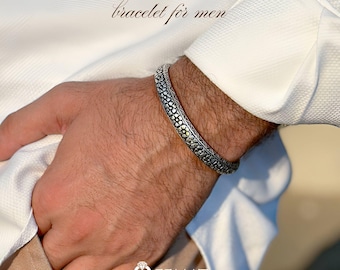 May include: A silver bracelet with a detailed floral pattern is worn on a wrist. The bracelet is on a person wearing a white shirt. The text "bracelet for men" is above the bracelet. The logo for Qeemat is in the bottom corner.