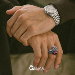 Può includere: Un anello d'argento a forma di drago con una gemma rossa su un dito. L'anello presenta dettagli intricati della testa e del corpo del drago. È visibile anche un orologio da polso argentato con cinturino in metallo.