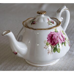 Op de afbeelding: Witte porseleinen theepot met een roze rozenbloemmotief en gouden details. De theepot heeft een gebogen tuit, een afgeronde body en een deksel met een goudkleurige knop. De handgreep is wit met gouden accenten. Een klassieker voor de thee.