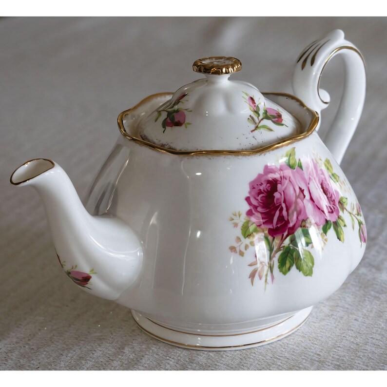 Op de afbeelding: Witte porseleinen theepot met een roze rozenbloemmotief en gouden details. De theepot heeft een gebogen tuit, een afgeronde body en een deksel met een goudkleurige knop. De handgreep is wit met gouden accenten. Een klassieker voor de thee.