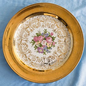 May include: A round ceramic plate featuring a wide, metallic gold rim with an etched pattern. The center is white, decorated with a central floral bouquet of pink roses, purple flowers, and yellow blossoms, surrounded by intricate gold filigree.