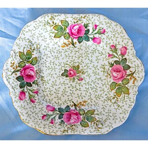 May include: A white, scalloped-edge serving tray with a floral design. The tray features pink roses, green leaves, and a delicate vine pattern. The rim is trimmed with gold.