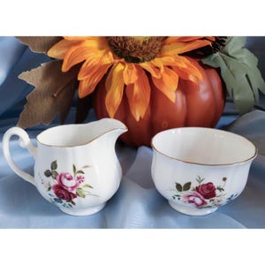 May include: A white porcelain creamer and sugar bowl set with a floral design featuring pink roses, green leaves, and blue accents. The set has a gold trim around the rim.