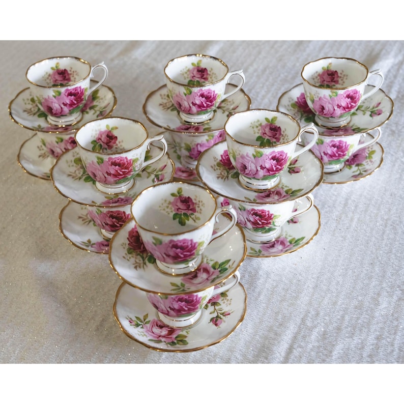 Op de afbeelding: Een set vintage theekopjes en schotels. Elk kopje en schotel heeft een witte achtergrond met roze rozenpatronen en gouden randen. De theekopjes zijn in een piramidevorm gerangschikt, wat de fijne details van het porselein benadrukt.