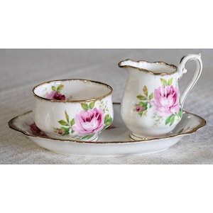 Op de afbeelding: Een vintage porseleinen theeservies met een suikerpot, een melkkan en een serveerschaal. De set is wit met roze rozenbloemmotieven en gouden randen. De schaal is ovaal en de melkkan heeft een gebogen handvat.