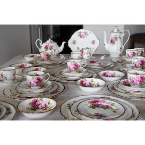 Op de afbeelding: Een complete vintage theeset met witte porseleinen stukken versierd met roze rozenbloempatronen en gouden randen. De set bevat theekopjes, schotels, borden, kommen, een theepot en een serveerschaal.