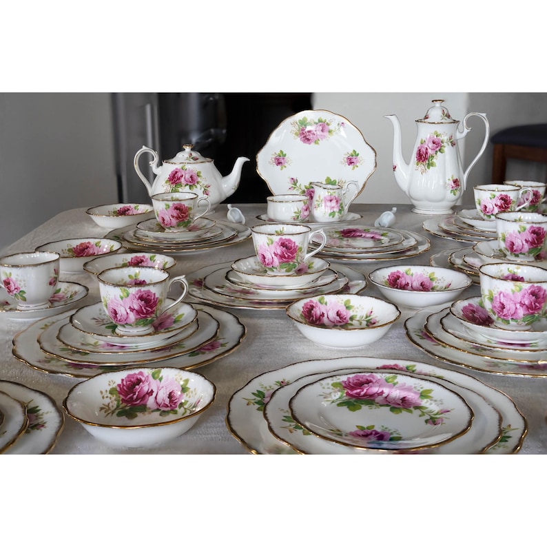 Op de afbeelding: Een complete vintage theeset met witte porseleinen stukken versierd met roze rozenbloempatronen en gouden randen. De set bevat theekopjes, schotels, borden, kommen, een theepot en een serveerschaal.