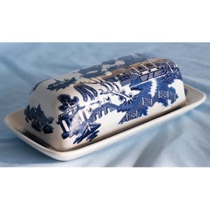 May include: A rectangular ceramic butter dish with a lid, featuring a white base and a blue willow pattern. The design depicts a landscape with a bridge, trees, and buildings. The lid has a curved top, and the base has a flat, raised edge.