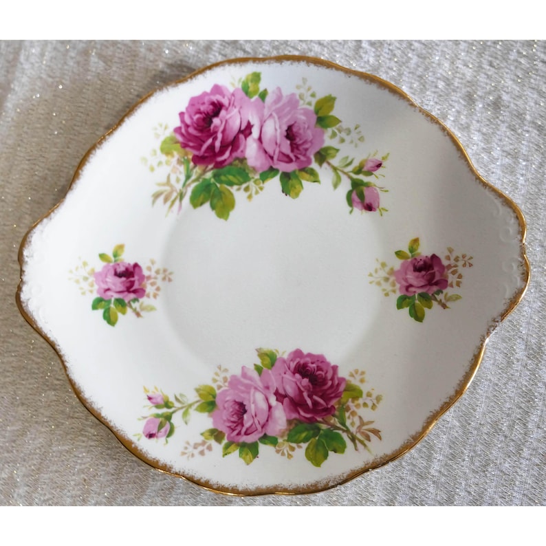 Op de afbeelding: Witte keramische schaal met een geschulpte rand en gouden rand. De schaal is versierd met roze rozen en groene bladeren. Een klassiek ontwerp voor het serveren van desserts of hapjes.