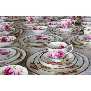 Op de afbeelding: Een verzameling vintage porseleinen theeserviezen, met kopjes, schotels, borden en kommen. Elk stuk is wit met een roze rozenbloemmotief en gouden rand. De sets zijn gerangschikt op een lichtgekleurd oppervlak.
