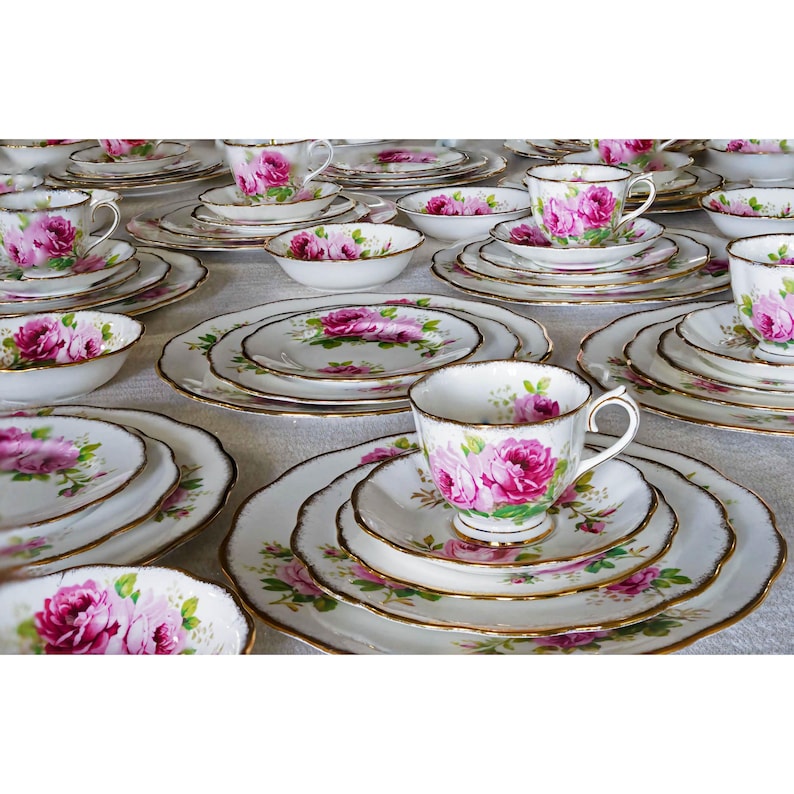 Op de afbeelding: Een verzameling vintage porseleinen theeserviezen, met kopjes, schotels, borden en kommen. Elk stuk is wit met een roze rozenbloemmotief en gouden rand. De sets zijn gerangschikt op een lichtgekleurd oppervlak.