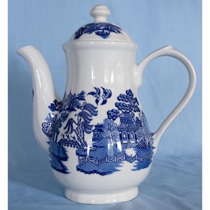 May include: A white ceramic teapot featuring a blue willow pattern. The design includes detailed illustrations of pagodas, bridges, trees, and birds. The teapot has a curved handle, a long spout, and a matching lid with a small knob on top.