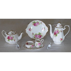 Op de afbeelding: Een wit porseleinen theeservies met roze rozenbloemmotieven en gouden randen. De set bevat een theepot, koffiepot, serveerschaal, melkkan, suikerpot en twee kleine lepels. Een klassieke theeservies.