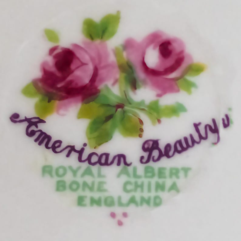 Op de afbeelding: Close-up van een wit porseleinen oppervlak met een bloemenontwerp. Twee roze rozen met groene bladeren zijn afgebeeld, met de woorden "American Beauty" in cursief. Daaronder staat de tekst "Royal Albert Bone China England."