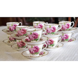 Op de afbeelding: Een verzameling vintage kopjes en schotels. Elk wit kopje en schotel is versierd met een roze bloemenpatroon en gouden rand. De kopjes zijn in een piramidevorm gerangschikt, wat een elegante presentatie cre&euml;ert.