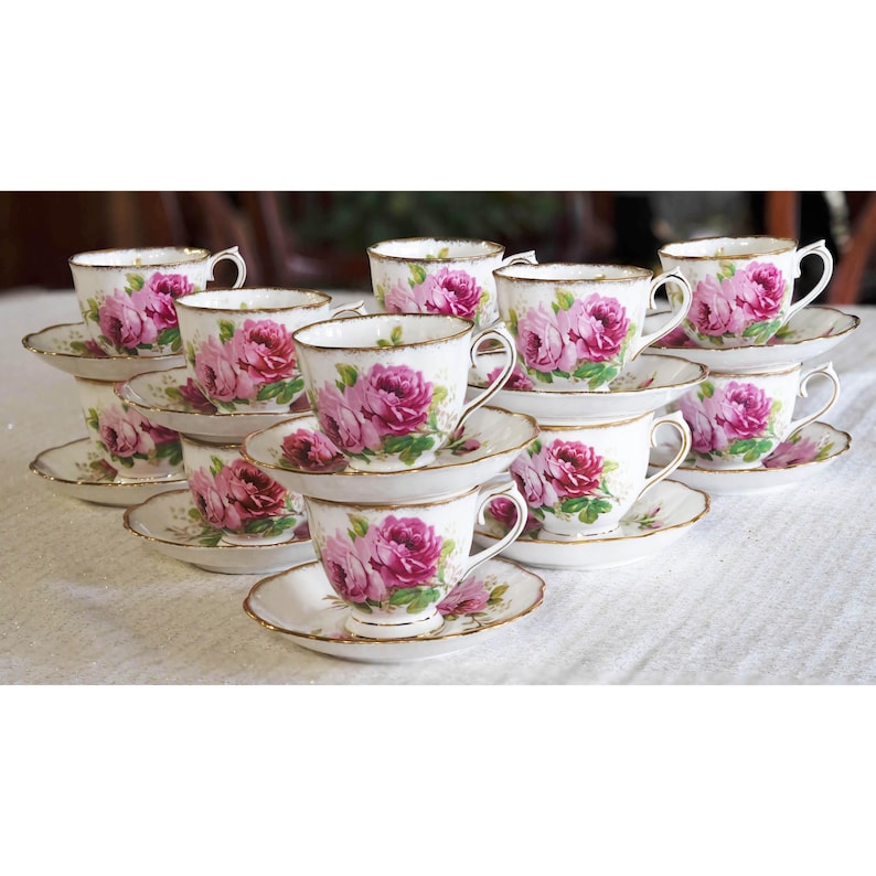 Op de afbeelding: Een verzameling vintage kopjes en schotels. Elk wit kopje en schotel is versierd met een roze bloemenpatroon en gouden rand. De kopjes zijn in een piramidevorm gerangschikt, wat een elegante presentatie cre&euml;ert.