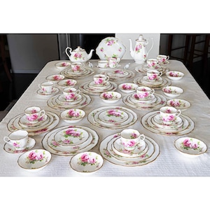 Op de afbeelding: Een complete vintage theeset met een rozenpatroon. De set bevat kopjes, schotels, borden, kommen, theepotten en een serveerschaal. Het porselein heeft een witte achtergrond met roze rozen en gouden randen, gerangschikt op een wit tafelkleed.