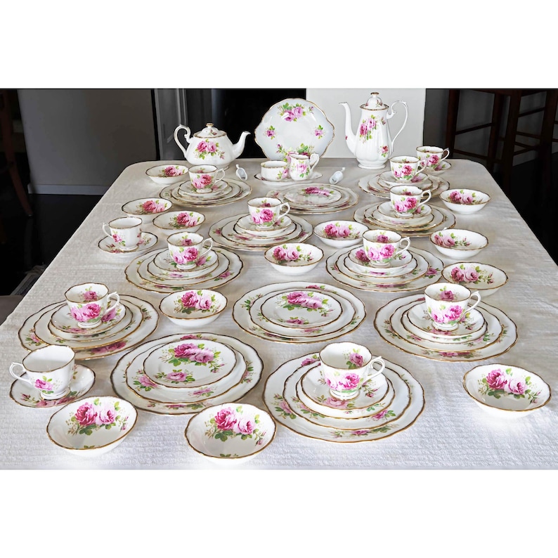 Op de afbeelding: Een complete vintage theeset met een rozenpatroon. De set bevat kopjes, schotels, borden, kommen, theepotten en een serveerschaal. Het porselein heeft een witte achtergrond met roze rozen en gouden randen, gerangschikt op een wit tafelkleed.