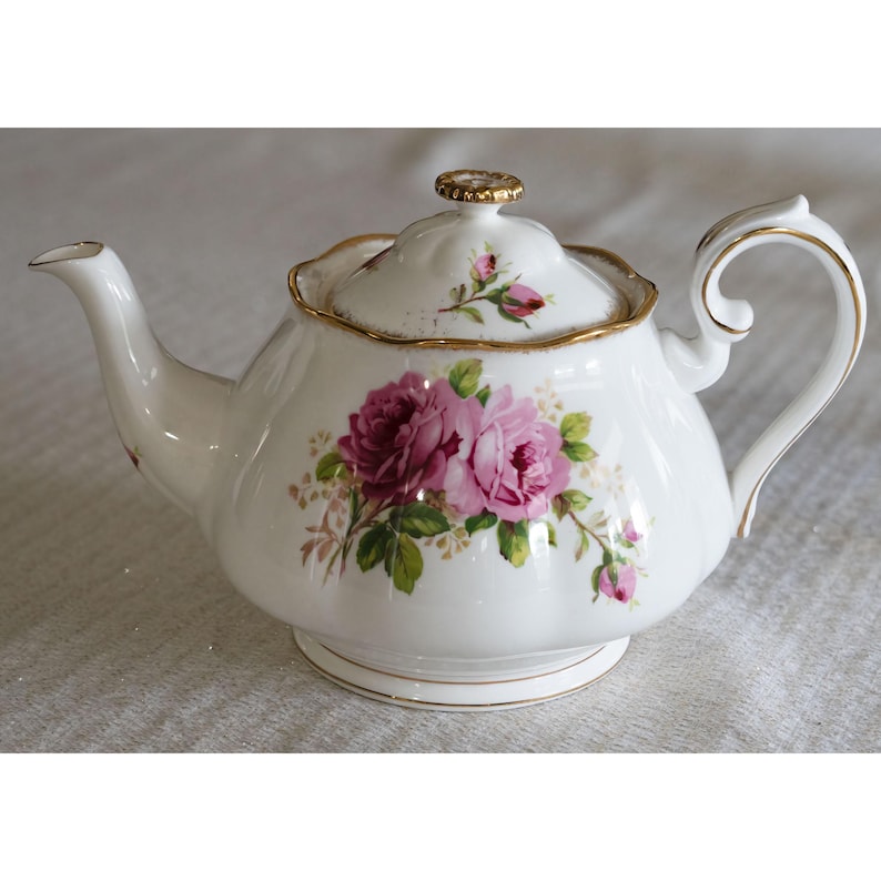 Op de afbeelding: Witte porseleinen theepot met deksel, versierd met roze rozen en groene bladeren. De theepot heeft een gouden rand op het deksel, de basis en het handvat. Een klassiek item voor een theeservies.