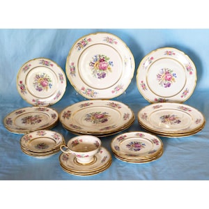 May include: A complete set of fine china with a floral design. The set includes dinner plates, salad plates, bowls, and a teacup with saucer. Each piece is white with a gold rim and a central floral arrangement in pink, purple, and blue.