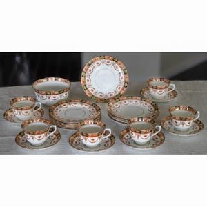 May include: A complete antique tea set featuring white porcelain cups, saucers, plates, and a bowl. The set is decorated with a repeating pattern of orange, green, and gold floral designs. The rims of the cups, saucers, and plates are trimmed with gold.