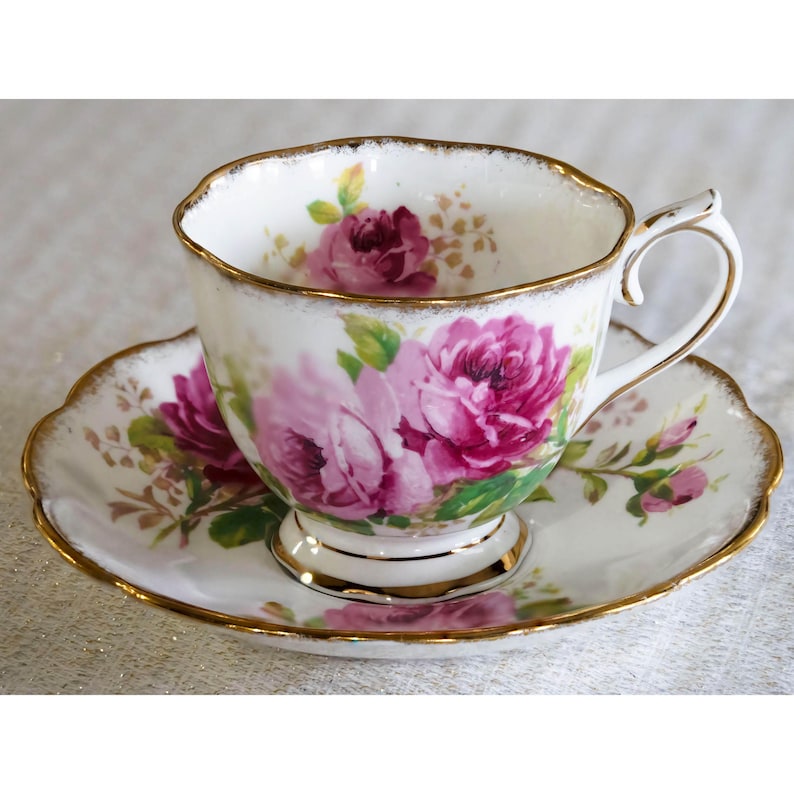 Op de afbeelding: Een witte porseleinen theekop en schotel met roze rozen en groene bladeren. De randen van de kop en schotel zijn goudkleurig. De theekop heeft een gebogen oor.