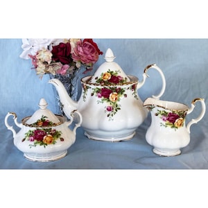 May include: A white porcelain tea set featuring a teapot, sugar bowl, and creamer. Each piece is adorned with a floral pattern of red and yellow roses, accented with gold trim. The set is displayed on a light blue surface.