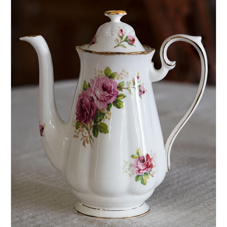 Op de afbeelding: Witte porseleinen koffiepot met deksel, versierd met een bloemenrozenpatroon in roze en groen. De pot heeft een gebogen handvat en een lange tuit. Gouden randen sieren de randen.