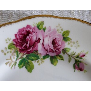 Op de afbeelding: Een witte porseleinen bord met een gouden rand, met een bloemmotief van roze rozen en groene bladeren. De rozen hebben verschillende tinten roze, van diep magenta tot lichtroze. Het bord heeft een geschulpte rand.