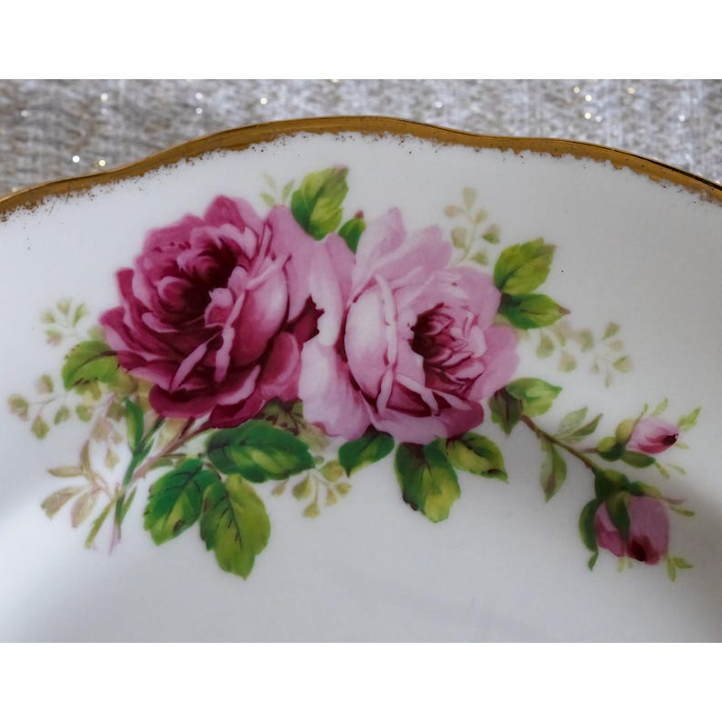 Op de afbeelding: Een witte porseleinen bord met een gouden rand, met een bloemmotief van roze rozen en groene bladeren. De rozen hebben verschillende tinten roze, van diep magenta tot lichtroze. Het bord heeft een geschulpte rand.