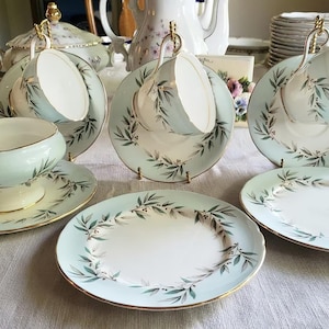 May include: A set of vintage teacups and saucers with a delicate green and gold floral design. The cups and saucers are arranged on a white tablecloth.