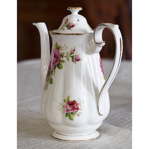 Op de afbeelding: Witte porseleinen koffiepot met een deksel, versierd met een bloemmotief van rozen in roze en rood. De pot heeft een gouden rand op het handvat, deksel en voet. Het handvat is gebogen en de tuit is slank.