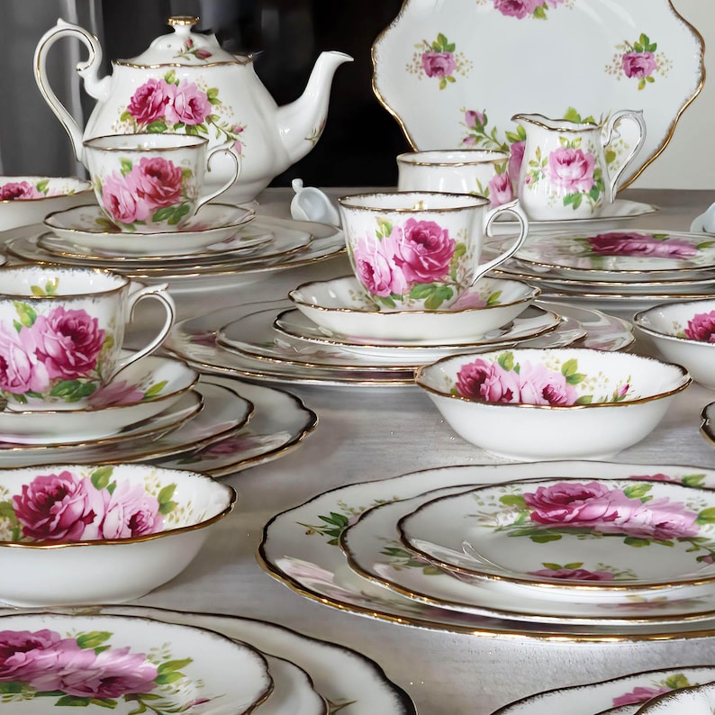 Op de afbeelding: Een wit porseleinen theeservies met een bloemmotief van rozen in roze en groen. De set bevat een theepot, kopjes, schotels, borden, kommen en een melkkan, allemaal met gouden rand. De opstelling staat op een wit oppervlak.