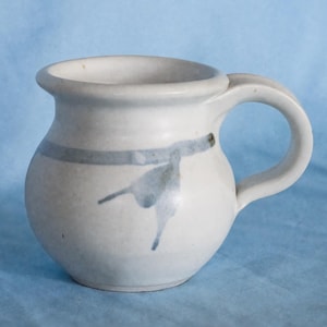 May include: A handmade ceramic mug with a rounded body and a curved handle. The mug is primarily white with a light blue band and abstract blue markings. The mug has a capacity of approximately 10 fluid ounces.