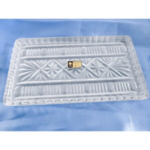 May include: A clear glass serving tray with a decorative etched pattern. The tray has a rectangular shape and a raised edge. The tray is made of glass and has a clear finish.